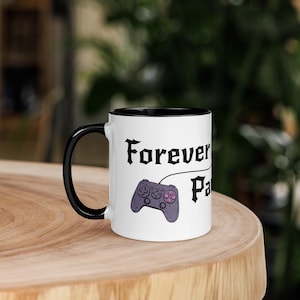 Könnte beinhalten: Ein weißer Keramikbecher mit schwarzem Henkel und schwarzem Innenrand. Der Becher zeigt eine schwarze Grafik eines Videospiel-Controllers, der durch eine Linie mit dem Text Forever Player verbunden ist, in einer fetten, stilisierten Schrift.