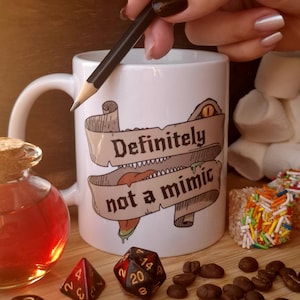 Può includere: Una tazza in ceramica bianca con la grafica di una pergamena recante il testo "Definitely not a mimic" e un occhio di mostro. La tazza è circondata da dadi poliedrici, chicchi di caffè, una piccola bottiglia di vetro con liquido rosso e marshmallow.