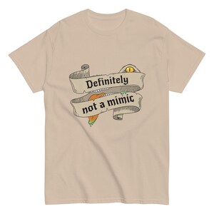 May include: A tan t-shirt featuring a banner design with the text "Definitely not a mimic." The banner has a cartoon eye and tongue design. The shirt is made of a soft material.