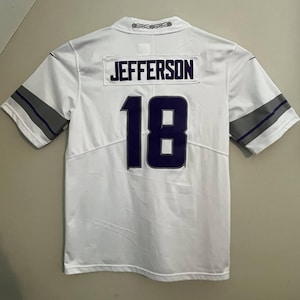 Op de afbeelding: Wit voetbalshirt met de naam "JEFFERSON" en het nummer "18" in marineblauw. Het shirt heeft grijs-witte strepen op de mouwen.