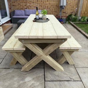 May include: A light-coloured wooden picnic table with attached benches. The table has an X-shaped base and is set on a stone patio. A bottle of wine, glasses, and a small plant are on the table.
