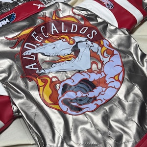 May include: Silver quilted jacket with red and white accents. The back features a circular emblem with the text "ALDECALDOS" surrounding a skull and flames design. The jacket has a quilted pattern and is made of a shiny material.