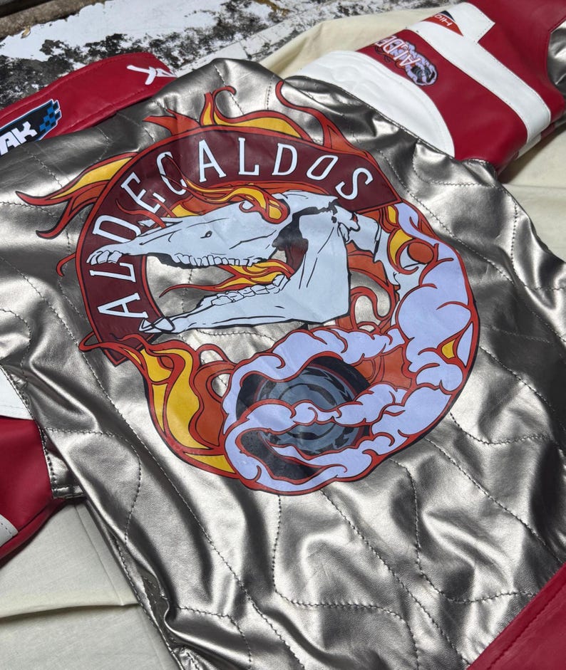 May include: Silver quilted jacket with red and white accents. The back features a circular emblem with the text "ALDECALDOS" surrounding a skull and flames design. The jacket has a quilted pattern and is made of a shiny material.