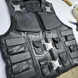 Puede incluir: Un chaleco t&aacute;ctico de cuero negro con el texto NovaHide, m&uacute;ltiples secciones acolchadas rectangulares y hebillas met&aacute;licas plateadas de liberaci&oacute;n r&aacute;pida en el pecho y los hombros. La prenda incluye varios bolsillos peque&ntilde;os con solapa.