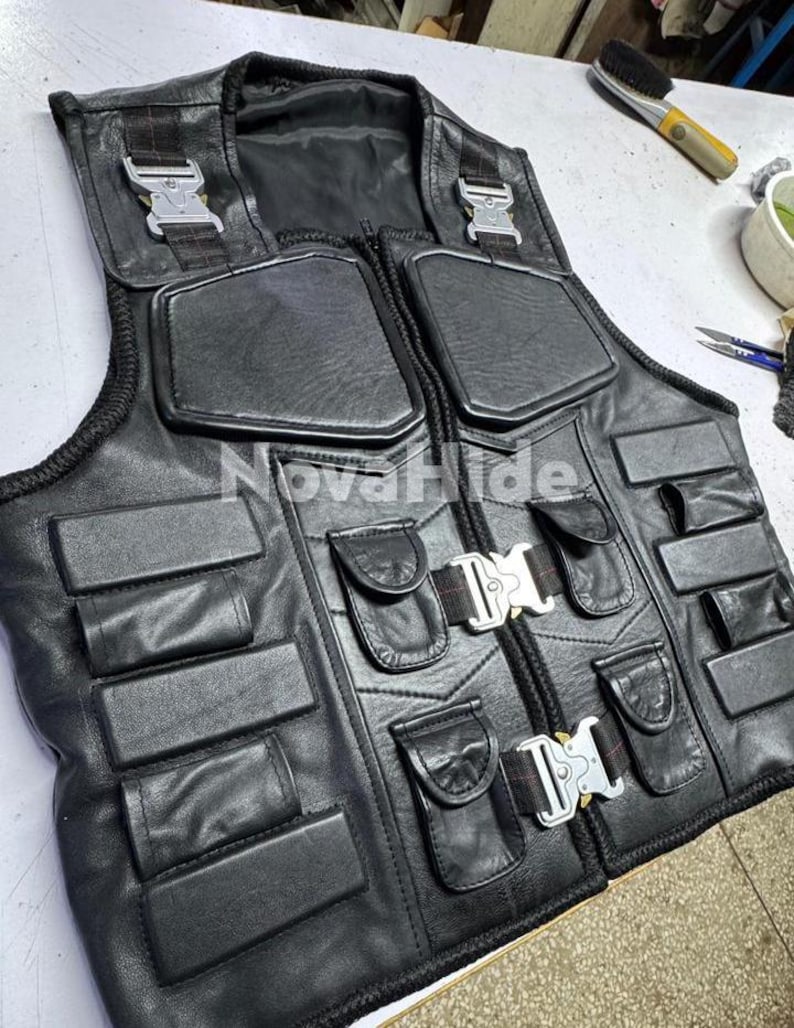 Puede incluir: Un chaleco t&aacute;ctico de cuero negro con el texto NovaHide, m&uacute;ltiples secciones acolchadas rectangulares y hebillas met&aacute;licas plateadas de liberaci&oacute;n r&aacute;pida en el pecho y los hombros. La prenda incluye varios bolsillos peque&ntilde;os con solapa.