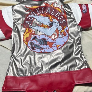 May include: Silver and red racing jacket with white accents. The back features a graphic with a skull, flames, and the text "ALDECALDOS." The jacket has "FUEL MY FIRE" printed on the bottom hem.
