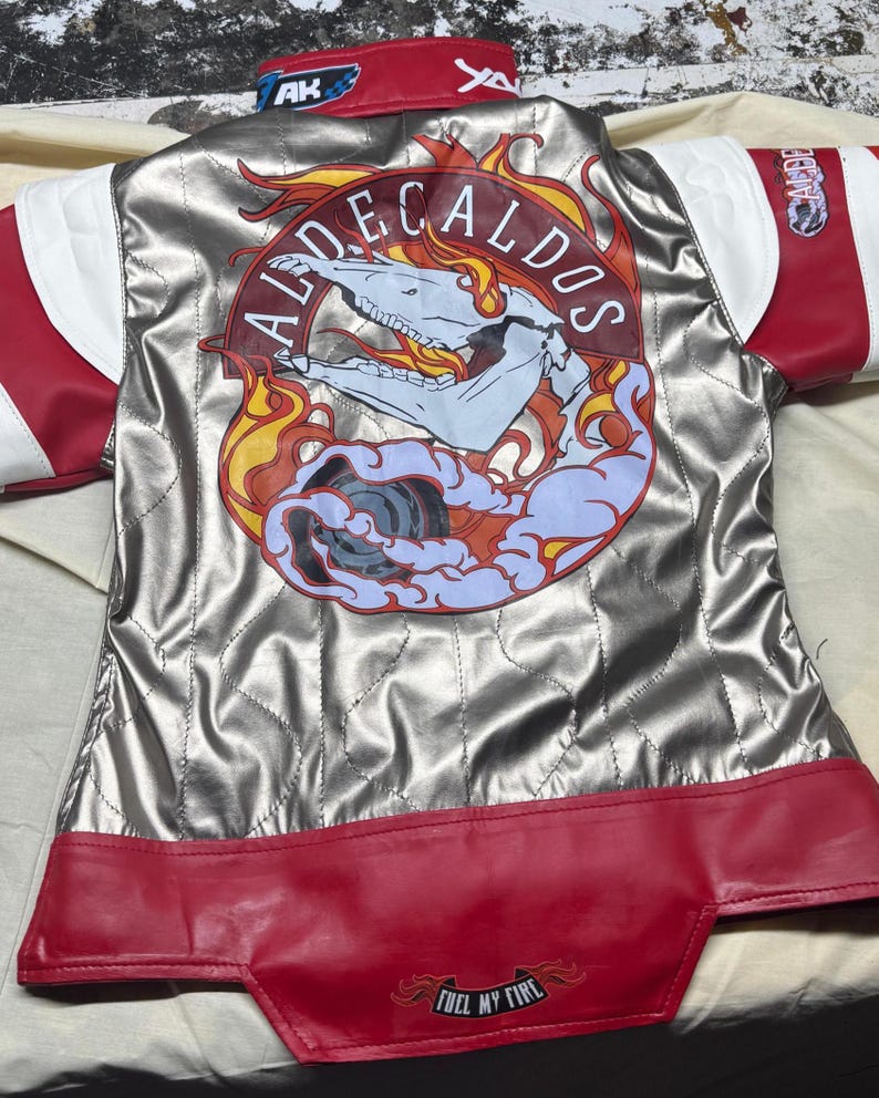 May include: Silver and red racing jacket with white accents. The back features a graphic with a skull, flames, and the text "ALDECALDOS." The jacket has "FUEL MY FIRE" printed on the bottom hem.