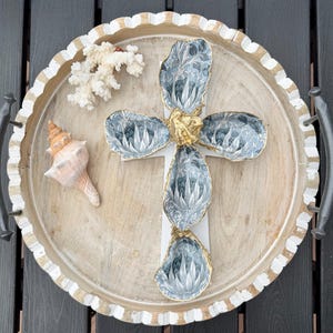 May include: A cross-shaped arrangement of four oyster shells with a blue and white floral pattern, featuring gold-painted edges and a central gold accent, placed on a round wooden tray with a white scalloped rim, alongside a white coral piece and a seashell.
