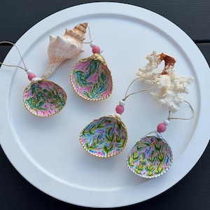 May include: Four decorative seashell ornaments featuring hand-painted green and blue botanical patterns on the interior. Each shell is accented with gold-painted edges, a pink and white bead, and a twine loop for hanging. One shell is attached to a spiral conch shell, and another is paired with a piece of white coral.