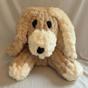 May include: A plush, handmade dog toy in shades of tan and cream. The dog has black eyes and nose, with cream-colored ears and paws. The soft, textured material gives it a cuddly appearance.