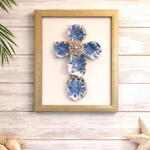 May include: Framed wall art featuring a cross design made from blue and white floral patterned ceramic pieces. A gold-colored frame surrounds the artwork, with a pearl and rhinestone brooch at the center. The background is a white wooden wall.