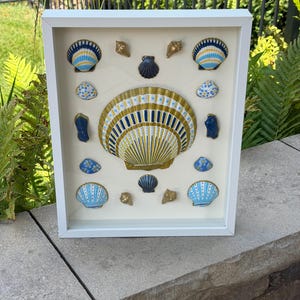 May include: A white shadow box frame containing a collection of seashells arranged on a cream background. The shells are painted in shades of blue, yellow, and gold with various patterns including stripes, dots, and floral motifs. A large central scallop shell is surrounded by smaller shells of different shapes and sizes.