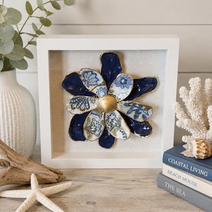 May include: A square white shadow box frame containing a three-dimensional flower sculpture crafted from layered oyster shells. The petals feature deep navy blue and intricate blue and white floral patterns with gold-painted edges and a gold center.