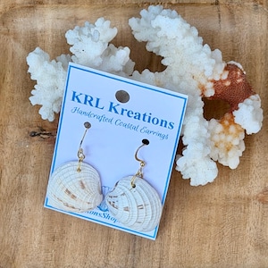 May include: A pair of white seashell earrings with gold-toned metal hooks attached to a white display card. The card features blue text that reads KRL Kreations Handcrafted Coastal Earrings. The earrings are positioned in front of white coral.