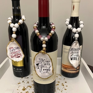May include: Three wine bottles with decorative beaded necklaces and shell-shaped tags. The tags have phrases like "Every empty bottle is filled with a great story" and "Wine a little, laugh a lot".