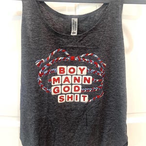 May include: Dark gray tank top with the words "BOY MANN GOD SHIT" in red and white block letters, surrounded by a braided design in red, white, and blue. The tank top is hanging on a gray hanger.