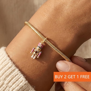 May include: A gold charm bracelet featuring a detailed nutcracker charm. The nutcracker is painted with pink, black, white, and gold accents. The bracelet is worn on a wrist. The image also contains the text "BUY 2 GET 1 FREE".