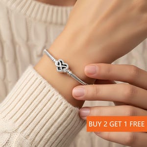 Könnte beinhalten: Ein silbernes Armband mit einem herzförmigen Anhänger, der ein Unendlichkeitssymbol und kleine, klare Steine ​​zeigt. Das Armband wird am Handgelenk getragen, und das Bild enthält den Text "BUY 2 GET 1 FREE" in einer orangefarbenen Box.
