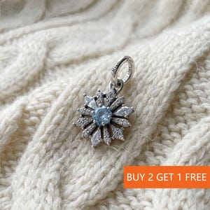 Könnte beinhalten: Ein silberner Charm mit Schneeflocken-Design. Der Charm hat einen hellblauen Mittelstein, der von klaren Kristallen umgeben ist. Der Charm liegt auf einer weißen Strickoberfläche. Der Text "BUY 2 GET 1 FREE" befindet sich in einem orangefarbenen Rechteck.