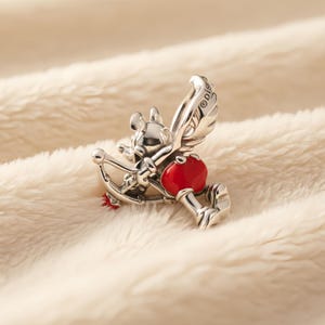 Puede incluir: Un charm de plata que representa un personaje de rat&oacute;n de dibujos animados con alas, sosteniendo un arco y una flecha roja en forma de coraz&oacute;n. El charm tiene un peque&ntilde;o lazo rojo y est&aacute; sobre un fondo suave de color crema.