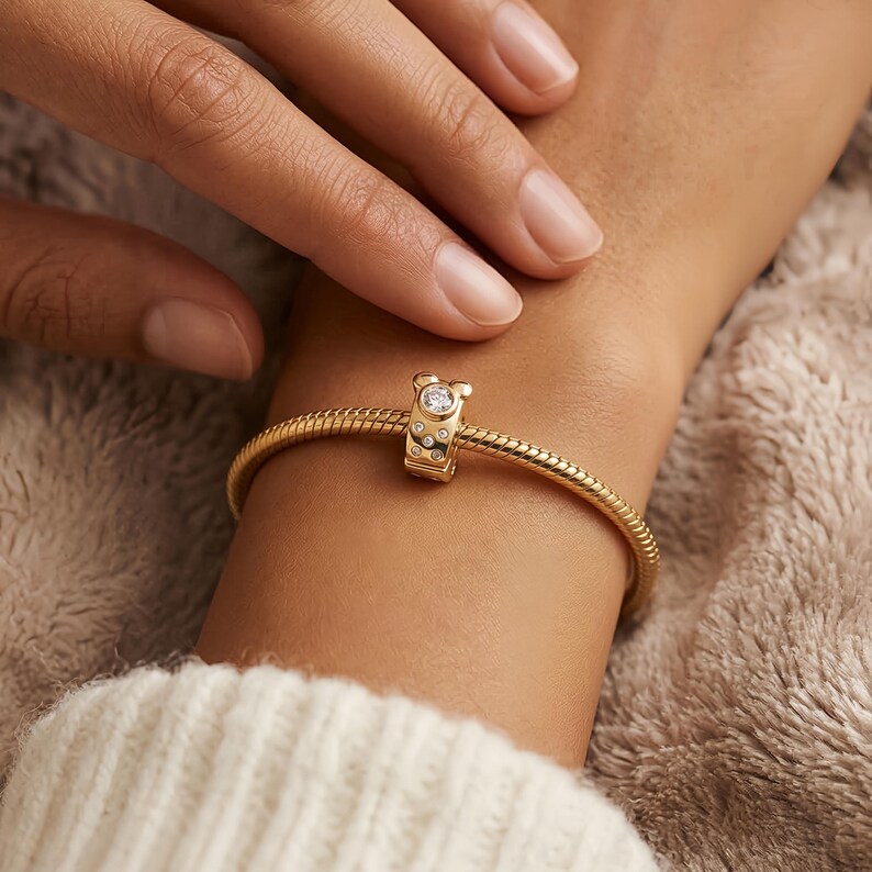 Puede incluir: Pulsera dorada con un charm. El charm presenta una gema transparente central y piedras m&aacute;s peque&ntilde;as alrededor. La pulsera tiene un dise&ntilde;o texturizado de cadena de serpiente. El charm tiene forma de oso. La pulsera se lleva en la mu&ntilde;eca.