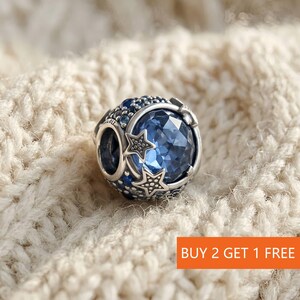Peut inclure: Un charm argenté avec un grand cristal bleu facetté au centre, entouré de petites pierres bleues et d'étoiles. Le charm est de forme ronde et posé sur un fond texturé crème. Le texte "BUY 2 GET 1 FREE" est visible en bas à droite.