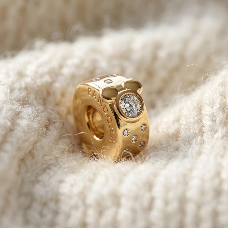 Puede incluir: Un charm dorado con un dise&ntilde;o inspirado en Mickey Mouse. Cuenta con una gema grande, redonda y transparente y varias gemas m&aacute;s peque&ntilde;as. La palabra "PANDORA" est&aacute; grabada en el lateral. El charm est&aacute; sobre un fondo blanco roto suave.