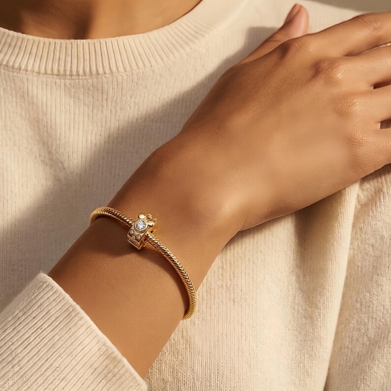 Puede incluir: Una pulsera dorada con una banda texturizada y un dije decorativo. El dije presenta una gema transparente central rodeada de piedras blancas m&aacute;s peque&ntilde;as. La pulsera se lleva en la mu&ntilde;eca, con un su&eacute;ter color crema en el fondo.