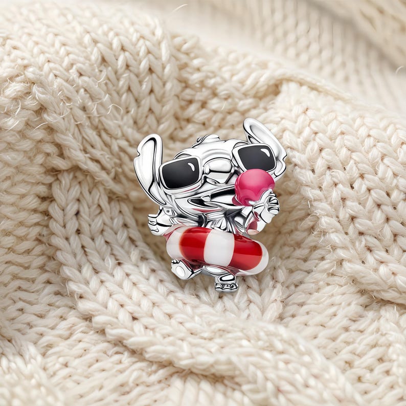 May include: A silver-toned metal charm shaped like a cartoon character wearing black sunglasses and holding a pink ice cream cone. The character is positioned inside a red and white striped circular float, featuring detailed engraved lines.