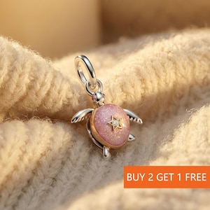 Può includere: Un charm a forma di tartaruga argentato e dorato con guscio rosa e design a forma di stella. Il charm ha un anello per attaccarlo a un braccialetto o una collana. Il testo "BUY 2 GET 1 FREE" è visibile.