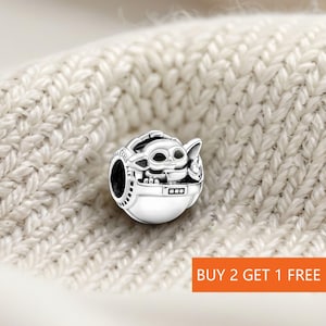 May include: A silver charm featuring a character from a popular science fiction series. The charm depicts a small, alien-like creature in a pod. The charm is set against a cream-colored, knitted background. The text "BUY 2 GET 1 FREE" is visible.