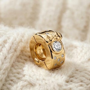 Puede incluir: Un charm dorado con un dise&ntilde;o de lazo y una gema grande, redonda y transparente. El charm est&aacute; adornado con varias gemas m&aacute;s peque&ntilde;as y transparentes. El charm est&aacute; sobre un fondo blanco suave y texturizado.