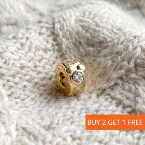 Puede incluir: Un charm dorado con una gema clara central y piedras claras m&aacute;s peque&ntilde;as. El charm tiene un dise&ntilde;o inspirado en Mickey Mouse y la palabra "PANDORA". El texto "BUY 2 GET 1 FREE" est&aacute; en un recuadro naranja.