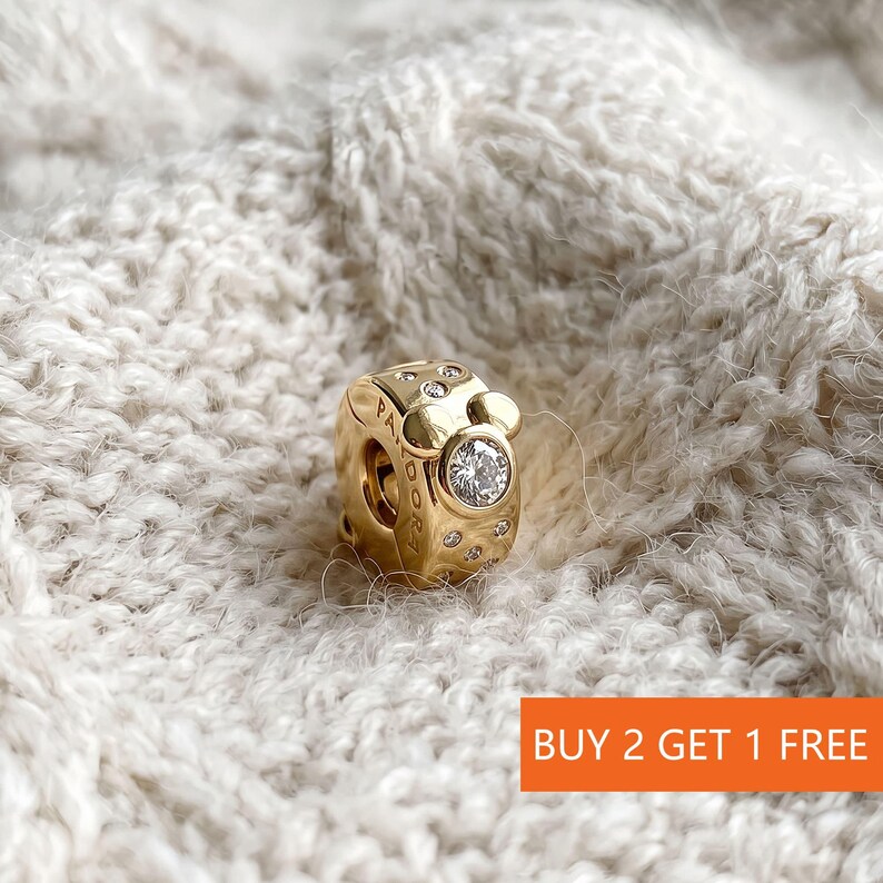 Puede incluir: Un charm dorado con una gema clara central y piedras claras m&aacute;s peque&ntilde;as. El charm tiene un dise&ntilde;o inspirado en Mickey Mouse y la palabra "PANDORA". El texto "BUY 2 GET 1 FREE" est&aacute; en un recuadro naranja.