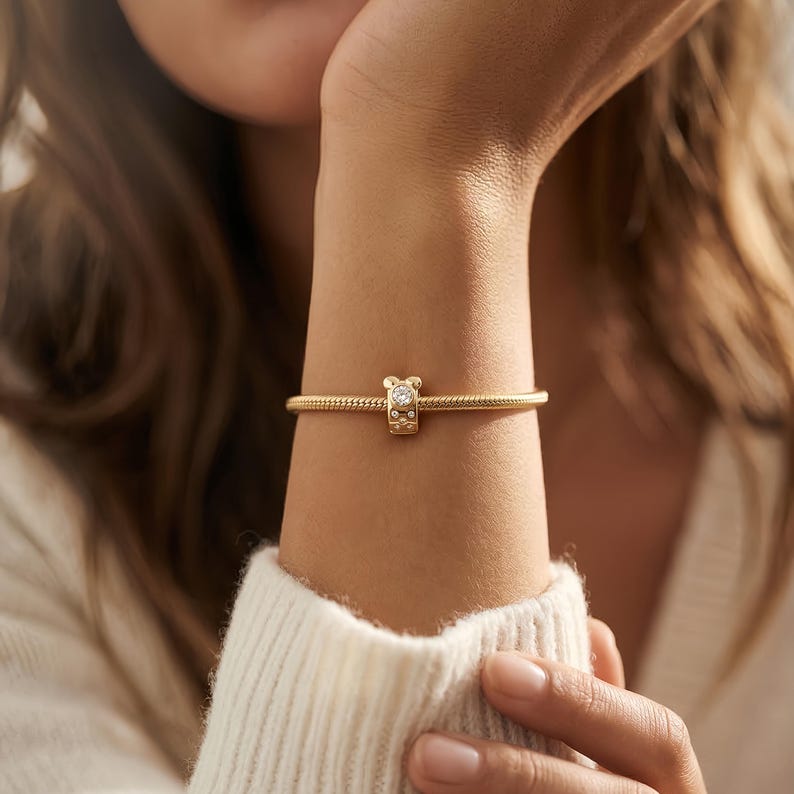 Puede incluir: Una pulsera dorada con un charm en forma de oso. El charm tiene una gema transparente y est&aacute; unido a una cadena dorada texturizada. La pulsera se lleva en la mu&ntilde;eca, con un su&eacute;ter blanco en el fondo. La cadena mide unos 18 cm.