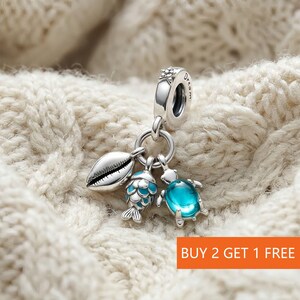 May include: A silver charm featuring a seashell, a fish with blue scales, and a turtle with a blue gem. The charm has a small floral design and the word "dream" engraved. Set against a white knitted background.