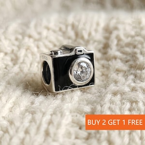 May include: A silver charm in the shape of a camera with a black enamel front and a clear, round gem for the lens. The charm is marked with "S925" and is set against a textured, off-white background. The charm is a jewelry item.