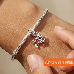 Puede incluir: Una pulsera de plata con un charm de Mickey Mouse en pose de Cupido, sosteniendo un arco y una flecha. El charm tiene detalles rojos y plateados. La pulsera est&aacute; en una mu&ntilde;eca. Texto en la imagen: BUY 2 GET 1 FREE.