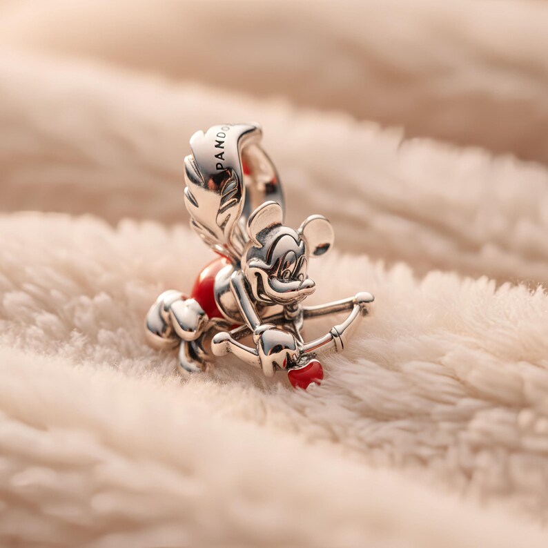 Puede incluir: Un charm plateado con un personaje de dibujos animados con arco y flecha, un coraz&oacute;n rojo y alas. El charm tiene la inscripci&oacute;n "PANDORA". El charm est&aacute; sobre un fondo blanco y suave.