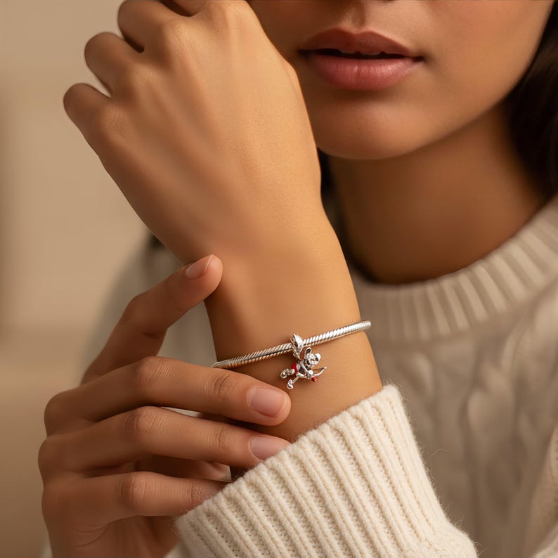 Puede incluir: Una pulsera de plata con un peque&ntilde;o y detallado dije. El dije presenta una peque&ntilde;a figura con detalles en rojo y blanco. La pulsera se lleva en la mu&ntilde;eca y el fondo est&aacute; borroso.