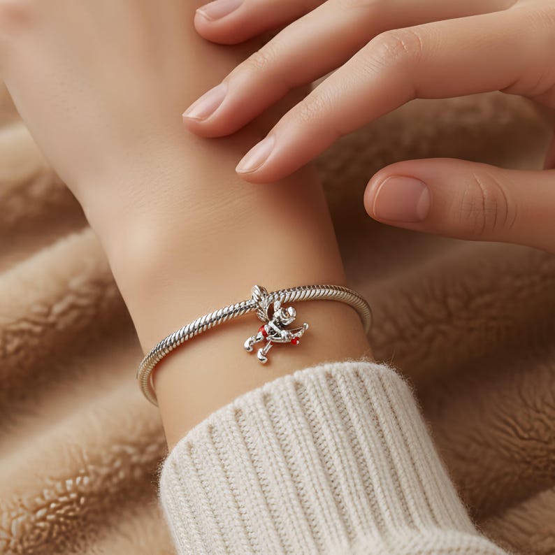 Puede incluir: Una pulsera plateada con un dije que representa un peque&ntilde;o Cupido con detalles rojos. La pulsera tiene un dise&ntilde;o de cadena de serpiente y se usa en la mu&ntilde;eca. El dije de Cupido sostiene un arco y una flecha.