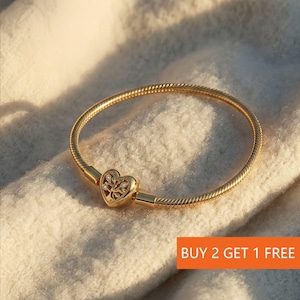 Könnte beinhalten: Ein goldfarbenes Armband mit herzförmigem Verschluss. Der Verschluss hat ein Baumdesign mit kleinen, klaren Steinen. Das Armband hat ein Schlangenketten-Design. Der Text "BUY 2 GET 1 FREE" ist im Bild zu sehen.