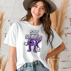 May include: White t-shirt featuring the text "SIX SEVEN" in a textured design above cartoon-like numbers. The numbers are purple with white gloves and shoes. The shirt is worn by a person.