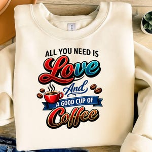 May include: Cream-colored sweatshirt with the text "ALL YOU NEED IS Love And A GOOD CUP OF Coffee." The design features a red coffee cup, coffee beans, and the word "Love" in red and blue.