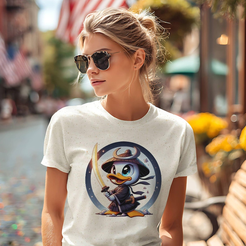 May include: A light-colored t-shirt featuring a cartoon duck dressed as a samurai, holding a sword. The duck is inside a blue circle with a starry background. The shirt is worn by a person wearing sunglasses.