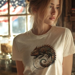 May include: Cream-colored t-shirt featuring a detailed illustration of a lizard with mechanical gears and intricate patterns in shades of brown, copper, and teal. The lizard's tail curls into a spiral design.