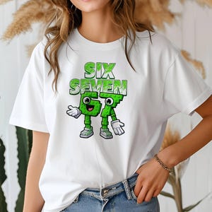 May include: White t-shirt featuring the text "SIX SEVEN" in green, above a cartoon graphic of the numbers 6 and 7 as green characters with arms, legs, and shoes. The shirt is made of a soft material.
