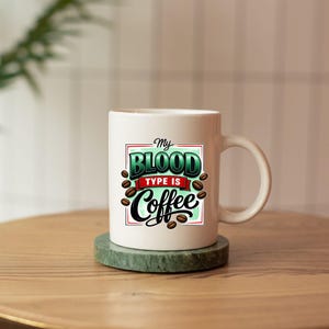 May include: White ceramic coffee mug with the text "My Blood Type Is Coffee" in a green, red, and black design. The mug features coffee bean illustrations and a curved handle. The mug sits on a green coaster.