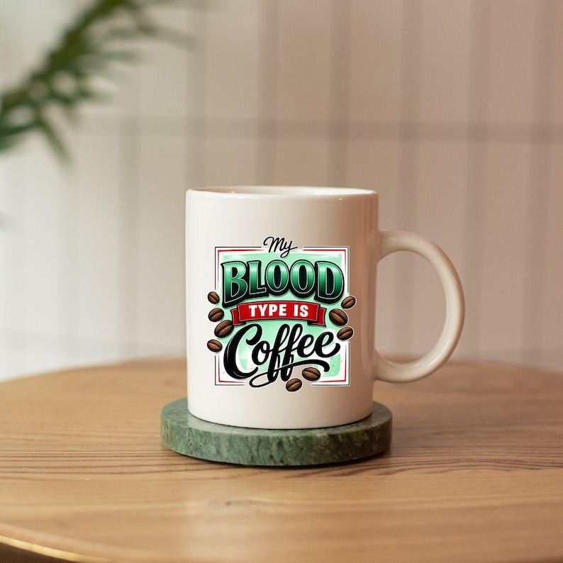 May include: White ceramic coffee mug with the text "My Blood Type Is Coffee" in a green, red, and black design. The mug features coffee bean illustrations and a curved handle. The mug sits on a green coaster.