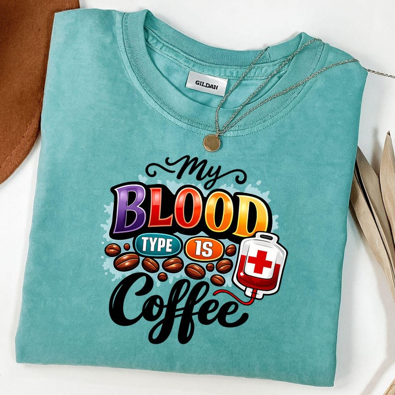 May include: A teal-colored t-shirt with the text "My Blood Type Is Coffee" in a colorful design. The graphic includes coffee beans and a blood bag. A gold necklace is draped across the shirt.